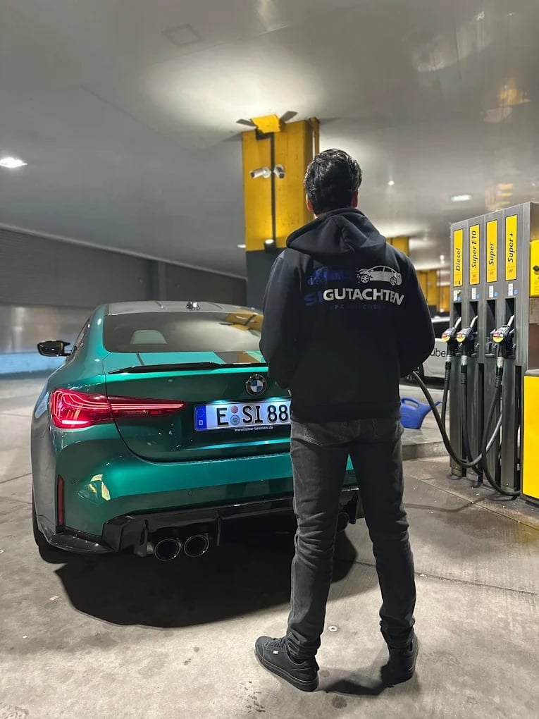 Person in dunkler Jacke steht an einer Tankstelle neben einem türkisfarbenen Fahrzeug